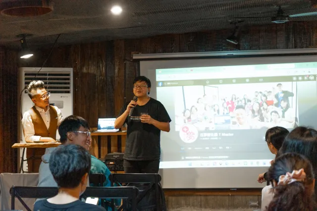 A speaker holds a microphone and presents to a seated indoor audience with a group photo on the projected screen behind him