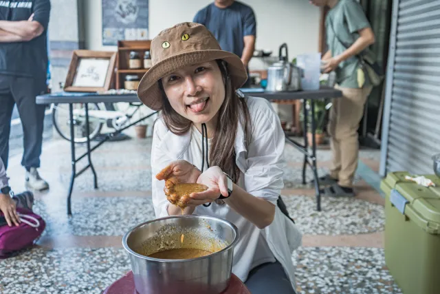 A smiling woman in a bucket hat playfully sticks out her tongue while handling food over a metal bowl at an outdoor cooking activity
