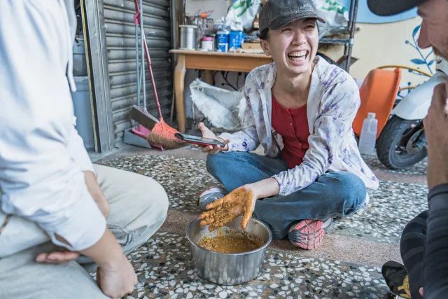 A laughing woman kneels on the ground mixing spices in a metal bowl with her hands