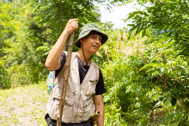 A guide carrying a wooden staff pauses on a lush forest trail, smiling and looking to the side
