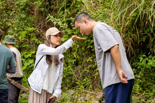 A woman in a white jacket gestures toward a man bending forward along a forest trail