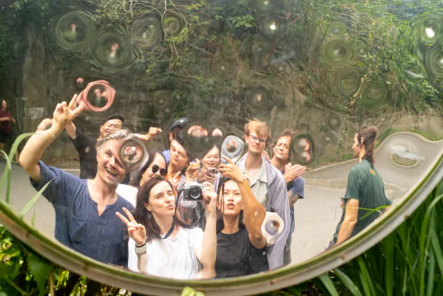 A group of tour participants pose and laugh while reflected in a large convex mirror outdoors