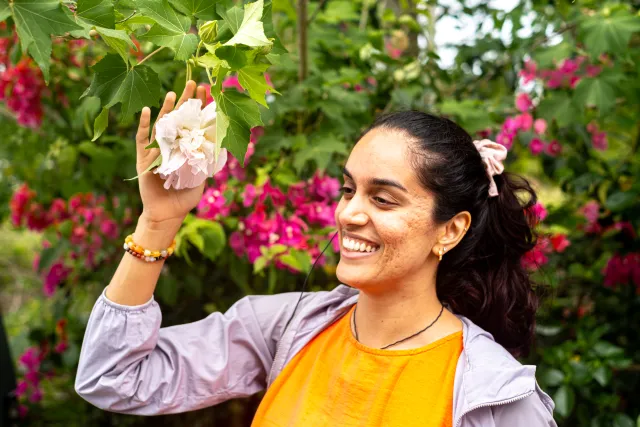 A smiling woman holds a white flower on a leafy branch amid pink blooms outdoors
