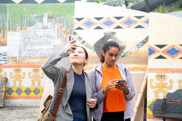 Two women photograph colorful Indigenous mosaic murals on an outdoor wall
