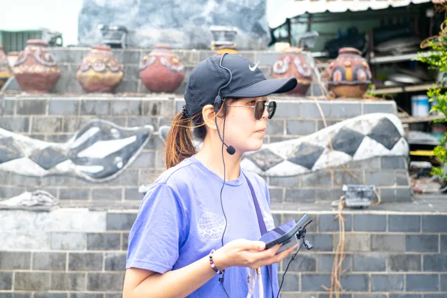 A woman with a headset microphone holds a device while guiding a tour outdoors near decorated ceramic urns