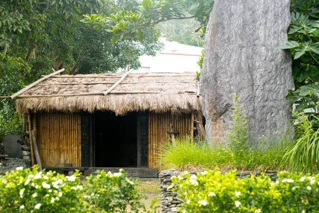 A traditional bamboo hut with thatched roof stands beside a large tree trunk amid lush greenery