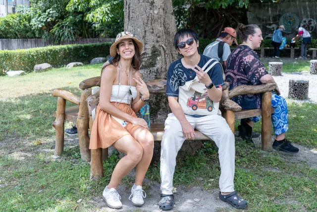 Two smiling visitors pose with peace signs on a wooden bench in a shaded park