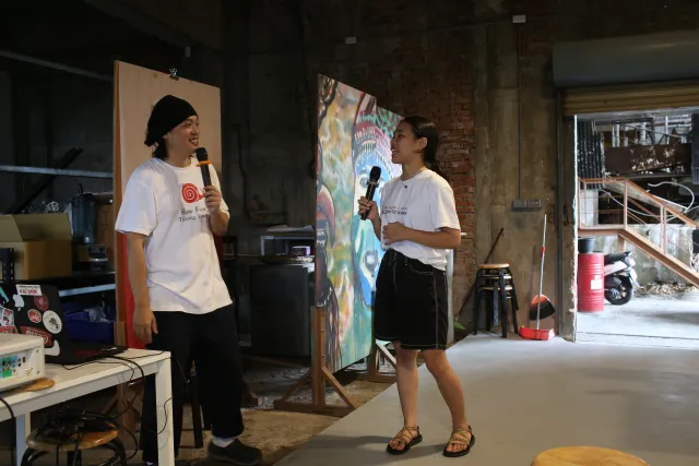 two hosts speaking into microphones inside an industrial venue with a colorful mural backdrop