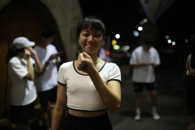 A young woman smiles at the camera on a night street, other participants in white T-shirts visible behind her