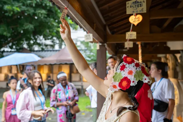 A woman in a floral crochet hat reaches up to hang a wooden wish plaque at a Japanese-style temple shrine
