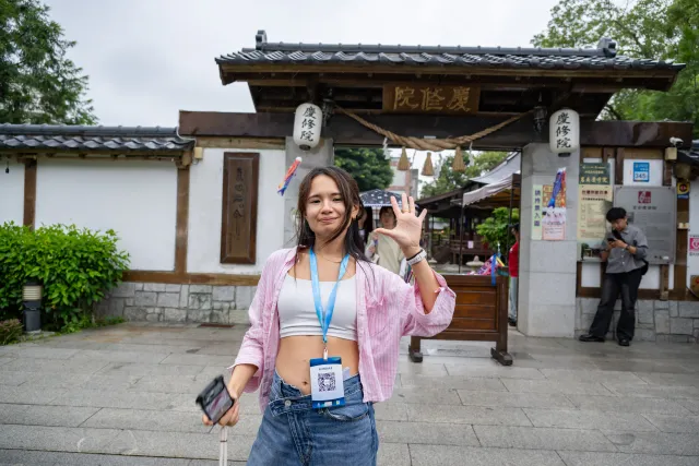 A young woman wearing a lanyard badge waves at the camera in front of Qingxiu Temple gate