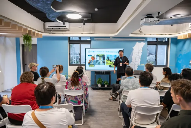 A presenter speaks to a seated audience of about twenty people in a modern blue-walled meeting room with a projected slide