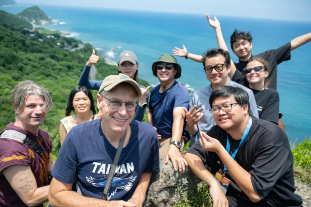 A cheerful group of eight people poses on a rocky hilltop overlooking the Pacific Ocean coastline