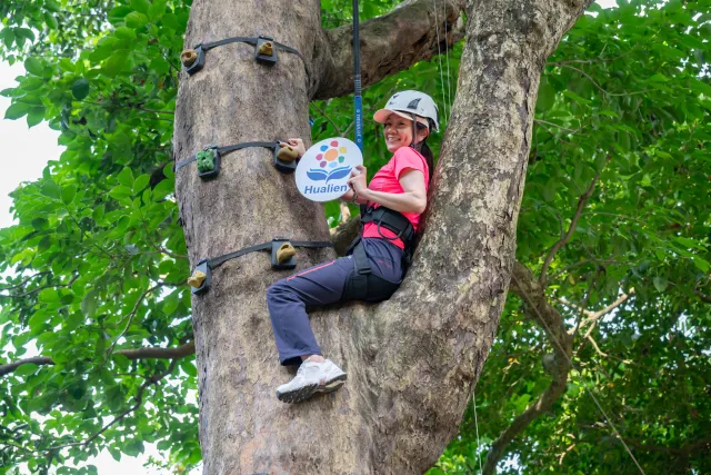 A smiling woman in a helmet clings to a large tree trunk while holding a Hualien sign