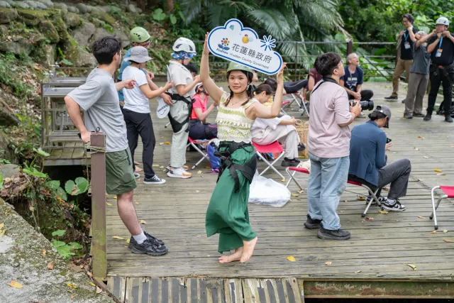 A barefoot woman in a green skirt holds up a Hualien County Government sign on a forest wooden deck, surrounded by onlookers and photographers