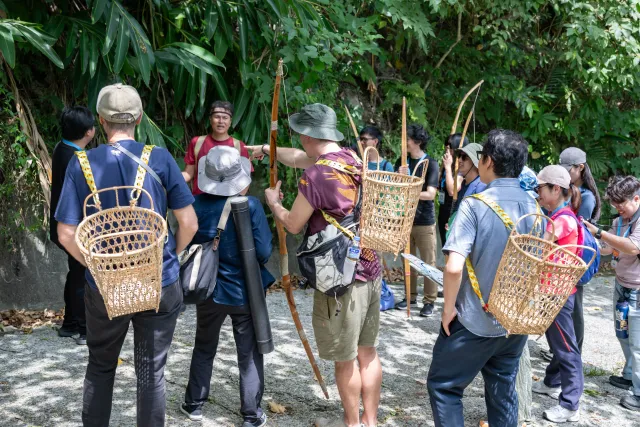Participants wearing woven backpack baskets hold traditional bows outdoors amid lush greenery