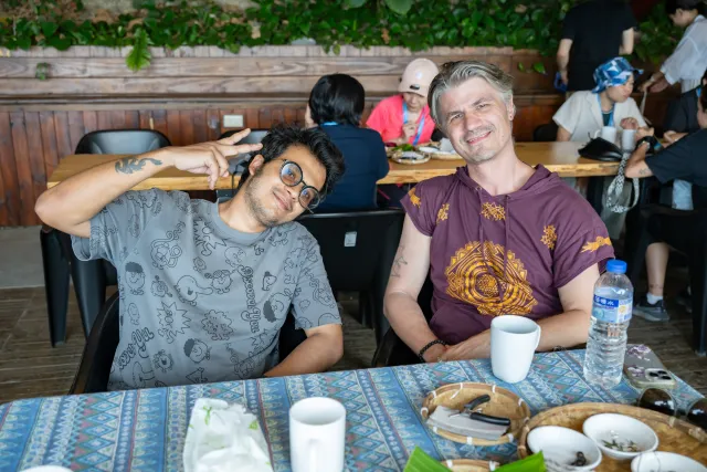 Two men smile and pose at a dining table in an open-air restaurant