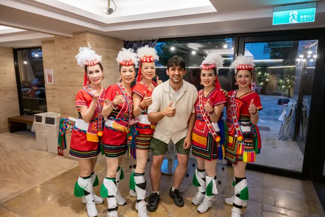 Five women in red Indigenous ceremonial dress pose with one man, all giving thumbs up indoors