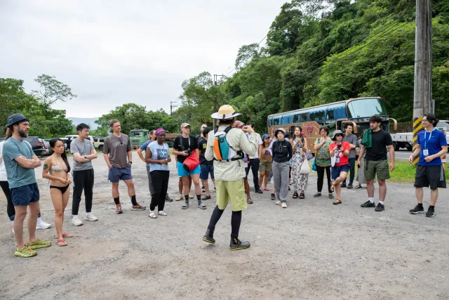 A guide addresses a group of participants gathered outdoors on a gravel lot near a forested hillside
