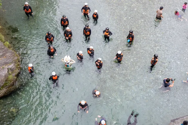 A large group of helmeted participants in life vests wading through a clear river, aerial view