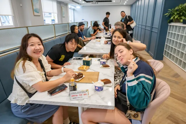 A group of visitors smile at the camera while seated at a long table with desserts and drinks in a bright café