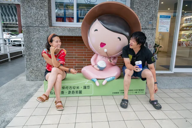 Two people sit on a bench beside a cheerful cartoon mascot statue outside a building