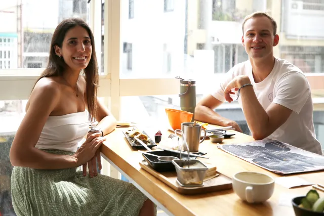 Two people smiling at a breakfast table by a bright window with food and drinks