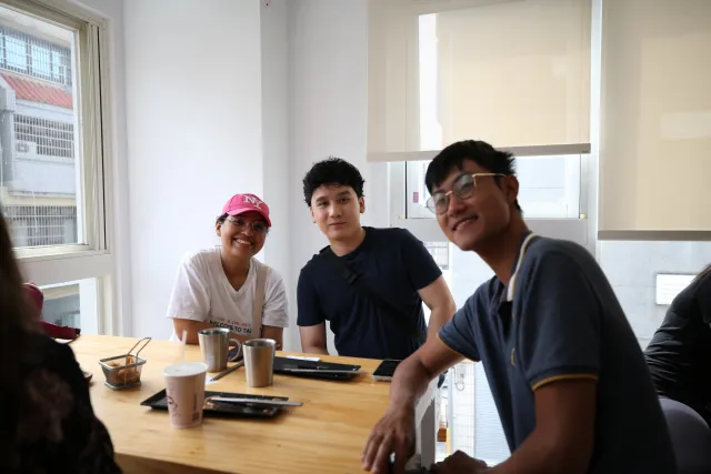 Three people smiling at a wooden table with coffee cups during a casual indoor breakfast