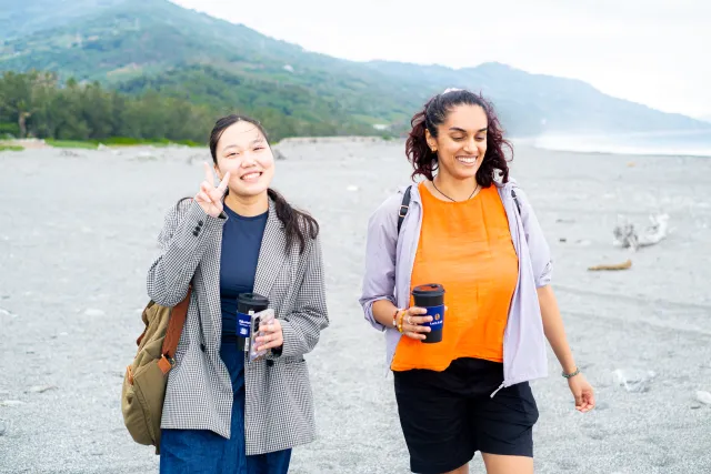 Two smiling women walk along a sandy beach holding travel mugs, one flashing a peace sign