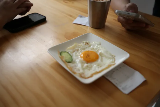 A fried egg with cucumber slices on a white plate on a wooden table, two people holding phones nearby