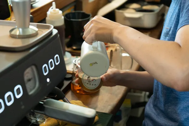 A person pours from a small ceramic pitcher near an espresso machine at a cafe counter