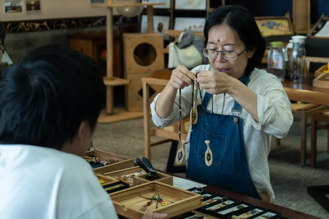 A craftsperson holds up a wooden pendant necklace to show a customer across a display table