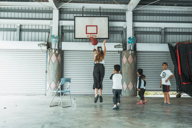 A woman jumps to shoot a basketball while three children watch in an open-air court
