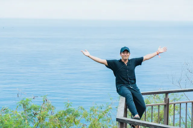 A man in a black cap sits on a railing with arms spread wide, smiling before a vast blue ocean
