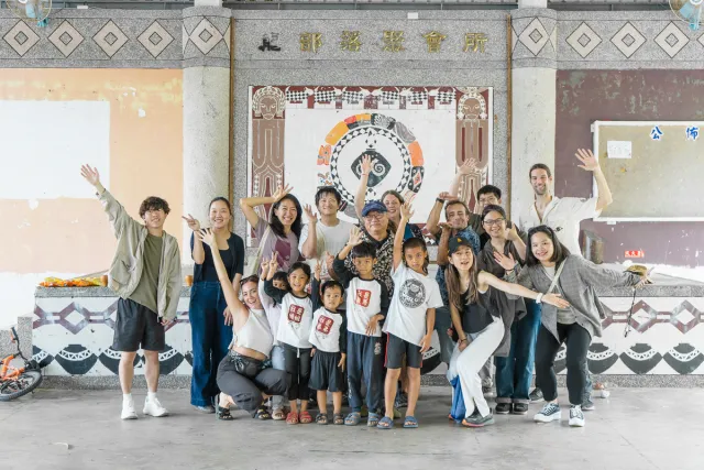 A group of adults and children pose cheerfully in front of an indigenous mural at a community hall