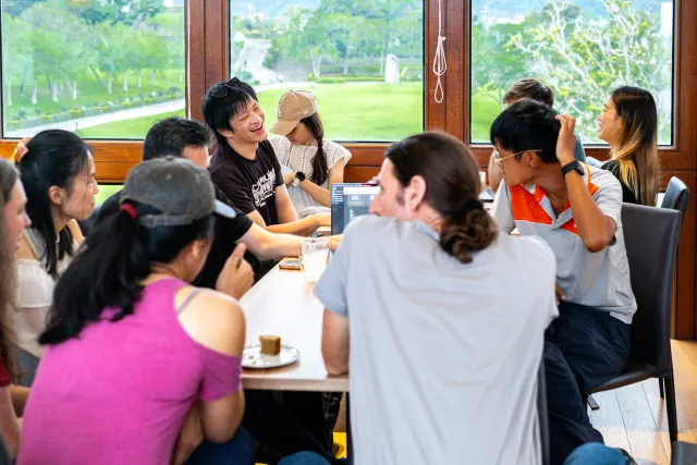 A group of people laugh and chat together around a long table in a bright cafe
