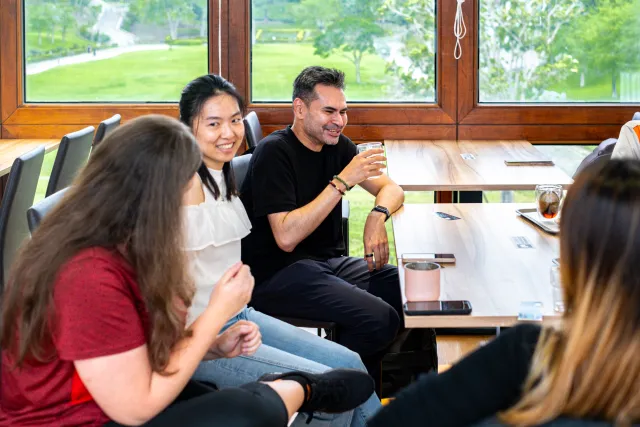 A man laughs and raises his drink while chatting with a small group at a wooden table by a window