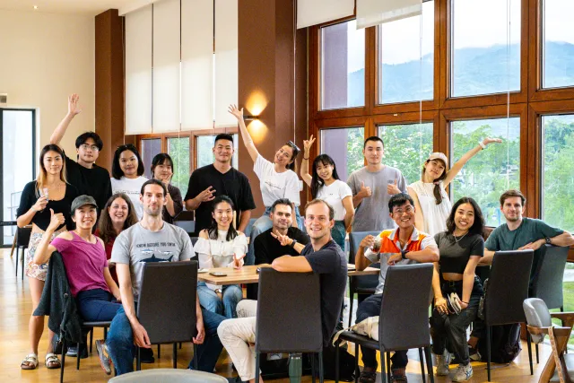 A large group of participants poses cheerfully around a table in a bright, airy venue with mountain views