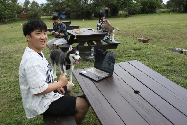 smiling man poses with a husky dog beside a picnic table on a grassy lawn