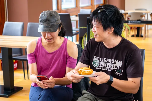 Two people laughing together indoors, one holding a phone and the other a pastry on a plate