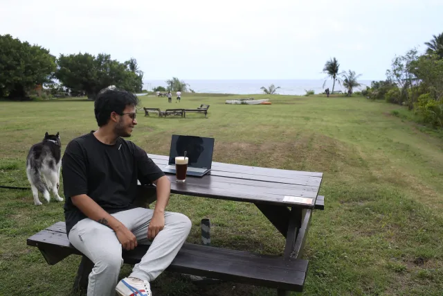 Man sitting alone at an outdoor picnic table with iced coffee and a laptop, a dog beside him, ocean in the background