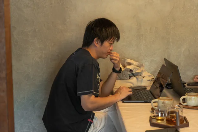 A young man sits at a café table, focused on his laptop with drinks nearby