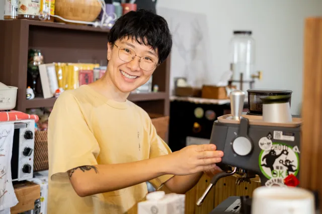 A smiling person in a yellow T-shirt operates an espresso machine at a coworking café counter
