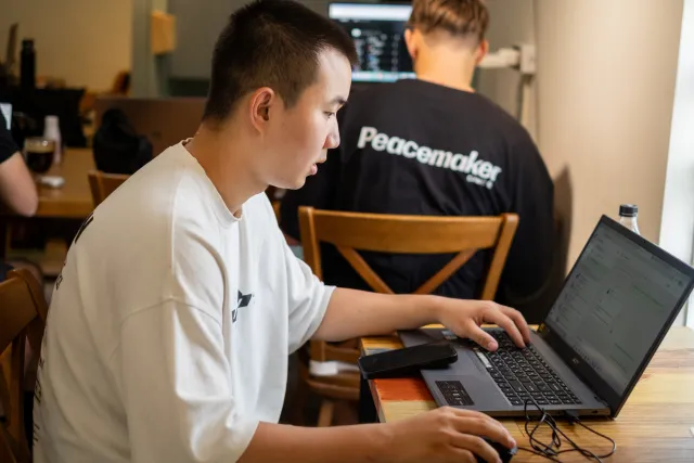 Young man typing on a laptop at a cafe table while others work nearby