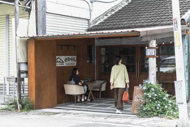 A person sits working on a laptop at an outdoor table while another walks into a wooden-facade café