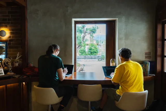 Two people working on laptops at a wooden bar facing an open window with a garden view