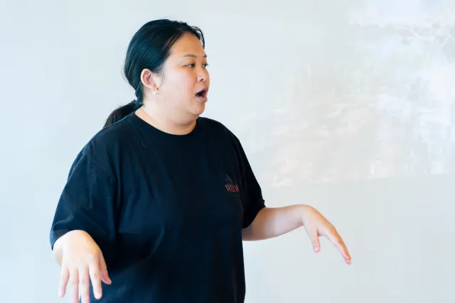 A woman in a black T-shirt speaks expressively with open hands in a bright room