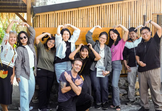 Group of ten participants posing cheerfully in front of a bamboo wall, making heart shapes with their hands
