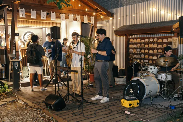 A jazz band performs outdoors at night on a wooden deck, with two saxophonists and a drummer playing under warm string lights