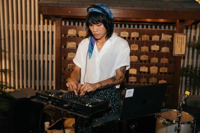 A tattooed woman in a white shirt and blue headband DJs at an indoor evening event